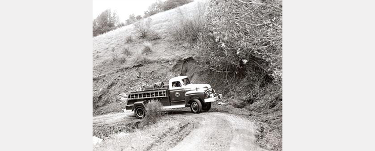 A fire truck drives up a curving road on the side of a hill, low brush in background. Black and white photo.