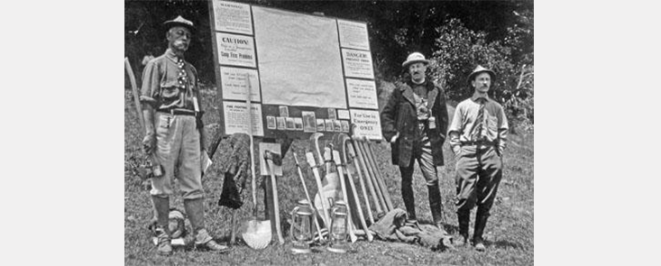 Three firefighters in front of an easel with shovels and other tools. On the easel are a map and posters. Black and white photo.
