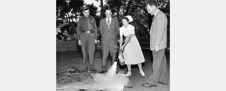 A woman in a nurse's uniform uses a fire extinguisher. Three men stand next to her, two in suits and one in a fire uniform. Black and white photo. 