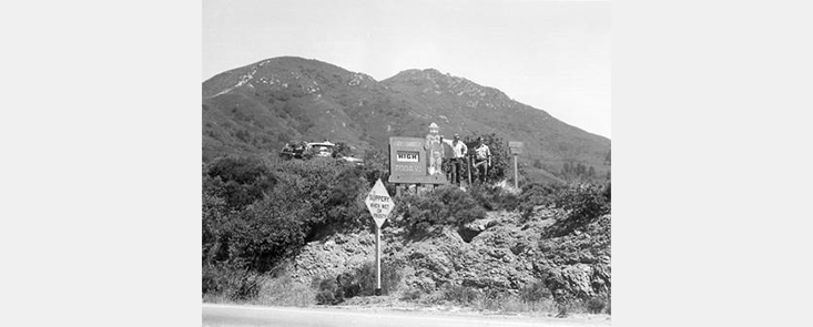 Two firefighters seen from a distance, standing next to a fire warning sign, mountains in background. Black and white photo.