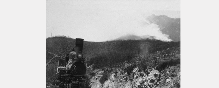 A steam train with mountains and smoke in background. Black and white photo.
