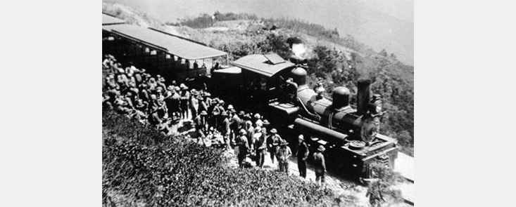 A large group of firefighters next to a steam train, on a hillside. Black and white photo.