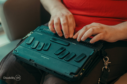 Humanware refreshable braille display sitting on the lap of person whose fingertips are on the Braille keys.
