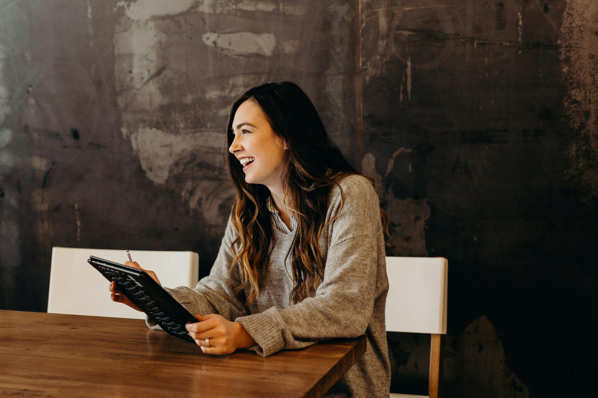 Person sitting at a table, smiling to the side while holding a tablet.