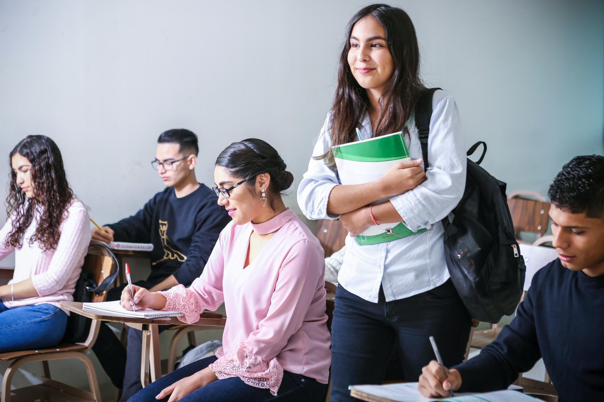 A group of young people in a college class, with one person walking between them holding books.