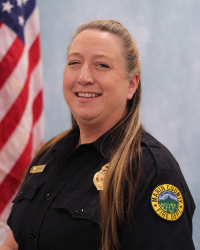 A headshot of a woman wearing a fire uniform in front of a blue background.
