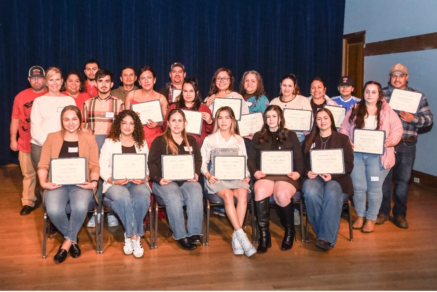 Group of approximately 20 people posing on a stage holding framed certificates at an awards or graduation ceremony.