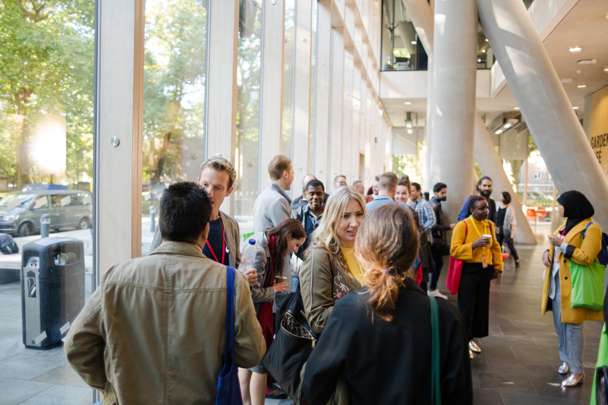 People gathered in a bright modern lobby, talking and mingling near large windows.