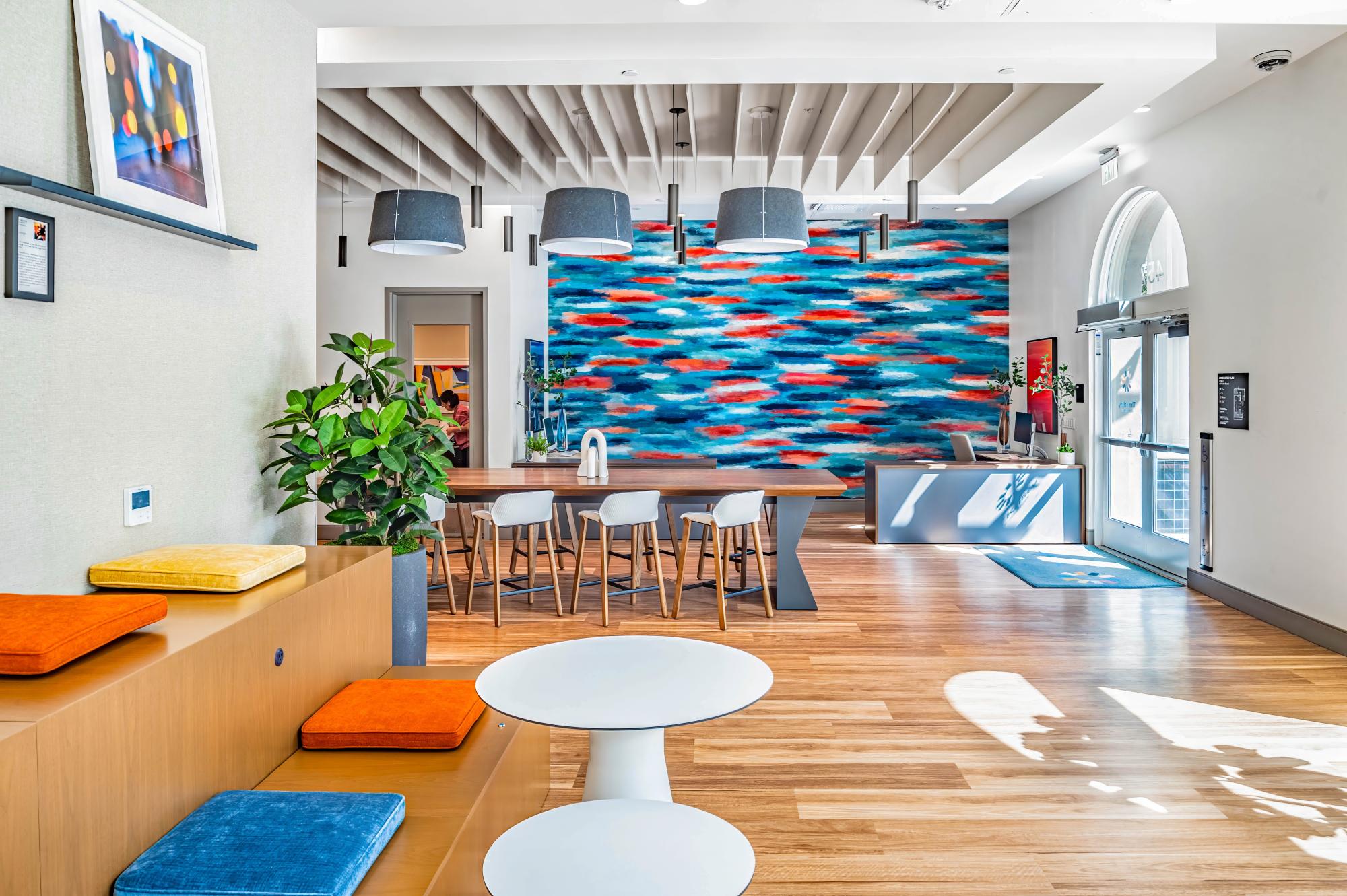 Sunlit common room with abstract blue and orange mural in the background, main door to the right and seating areas.