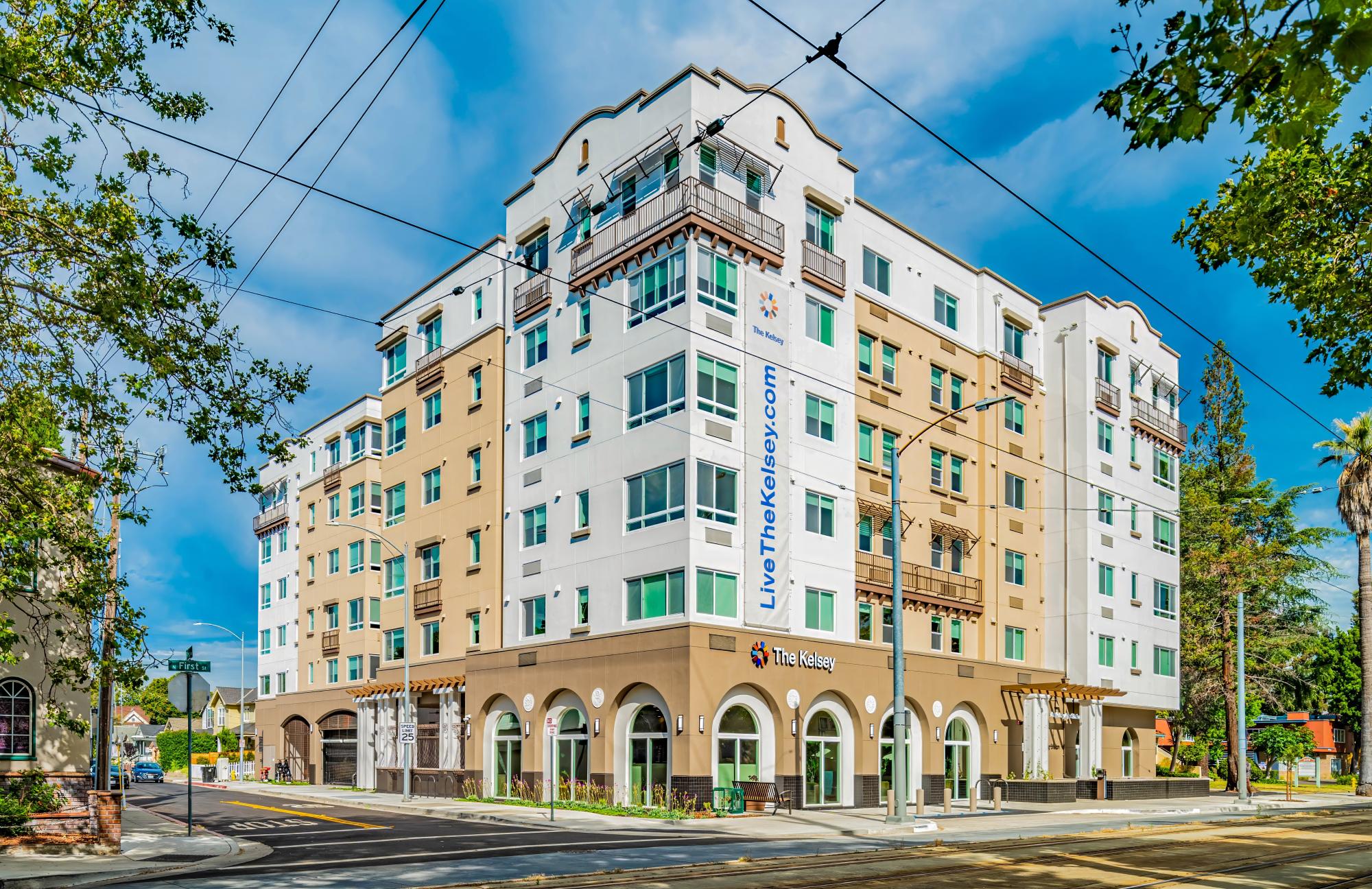 Exterior view of a modern multi-story apartment building with a tan and white facade, large arched windows on the ground floor, and a tall vertical banner that reads "LiveTheKelsey.com".